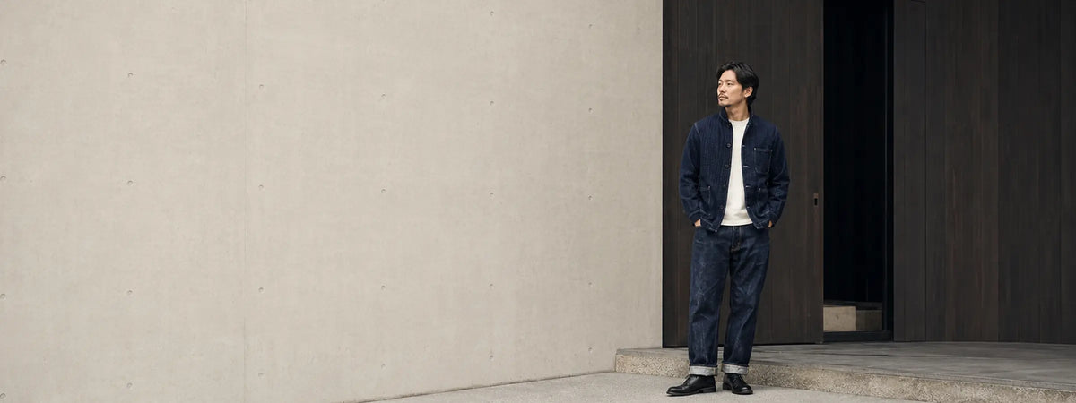 Homme vêtu d’une veste en indigo texturé et d’un jean japonais devant une architecture minimaliste en béton et bois.
