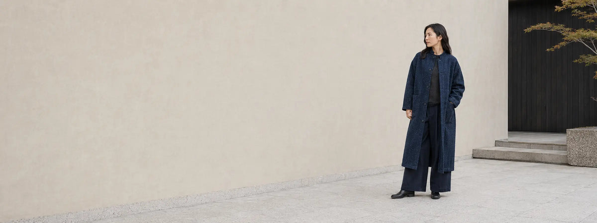 Femme portant un long manteau japonais en indigo texturé devant une architecture minérale épurée.