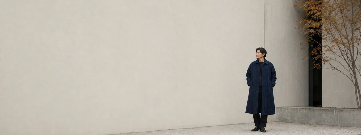 Homme portant un long manteau indigo japonais texturé devant une façade minérale épurée.